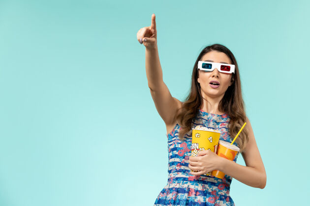 抱着正面图年轻的女性拿着爆米花 戴着d墨镜在蓝色的表面上喝酒前面爆米花电影院