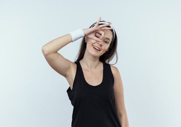 头带戴着头巾的年轻女健身师正在做“ok”手势 手指穿过这个手势 微笑着站在白色的墙上通过微笑健康