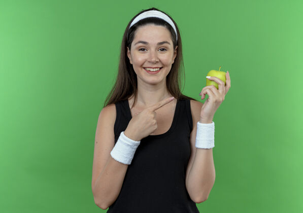 欢呼戴着头巾的年轻健身女 手拿青苹果 手指着苹果 站在绿色的墙上 开心地笑着站着年轻微笑