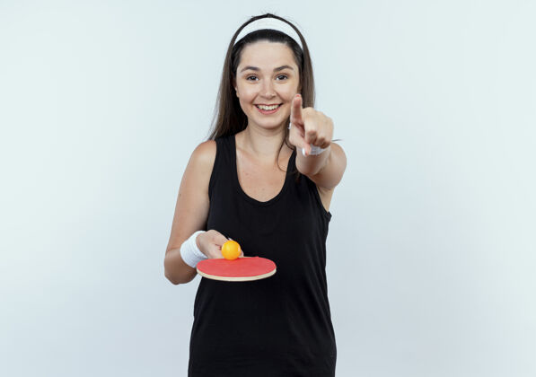 球拍戴着头巾的年轻健身女士手持球拍和乒乓球 手指着手指 站在白色的墙上欢快地笑着乒乓球欢呼抱着