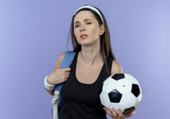 年轻戴着头巾 背着背包 拿着足球的年轻健身女士站在蓝色的墙上 看起来很困惑站抱背包