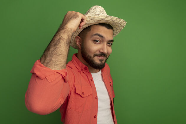 男人年轻的留着胡子的男人穿着橘色衬衫 戴着夏天的帽子 站在绿色的墙壁上 面带微笑 充满自信夏天胡须自信