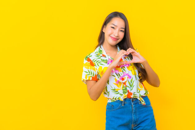 女性美丽的亚洲年轻女子的肖像 穿着五颜六色的衬衫 在黄色的墙上做心形标志享受微笑人