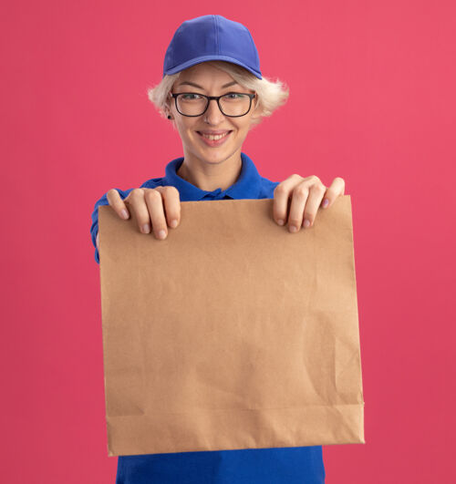 眼镜身穿蓝色制服 头戴眼镜 手拿纸包 面带微笑的年轻送货员在粉红色的墙上帽子送货微笑