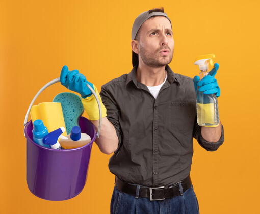 清洁年轻人穿着便服 戴着帽子 手里拿着水桶 手里拿着清洁工具 站在橙色的墙上困惑地看着一边拼图水桶靠边站