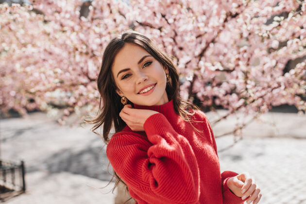 健康樱花附近穿着红色毛衣的美丽女孩的肖像身着现金宝石套装的迷人女人微笑着看着花园里的相机年轻华丽咖啡