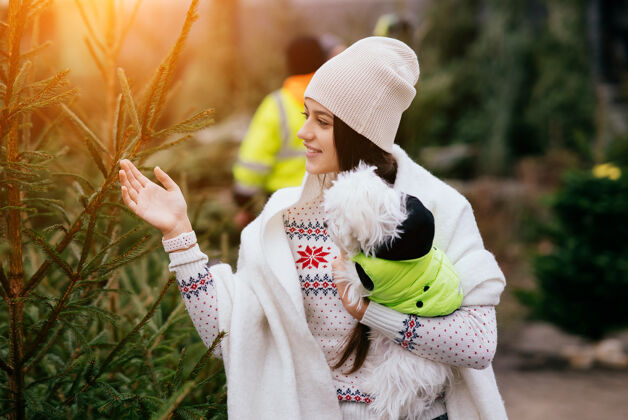 美丽市场上一棵绿色的圣诞树旁 一个抱着一只白色狗的女人约克郡主人朋友