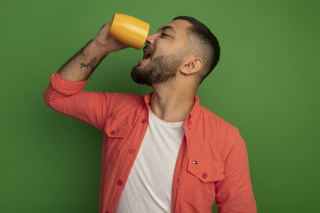 喝酒身穿橘色衬衫的留着胡子的年轻人站在绿色的墙上喝茶年轻人男人胡子