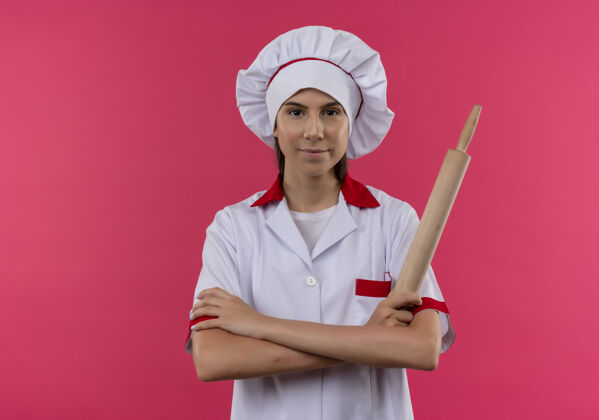 年轻年轻自信的高加索厨师女孩身着厨师制服 双手交叉握着擀面杖 被隔离在粉色空间和复印空间空间卷烹饪
