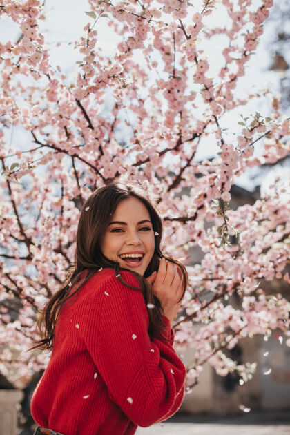 自然穿着鲜艳毛衣的迷人女人对着盛开的樱花笑穿着红色套装的酷黑发女人微笑着享受春天温柔芳香花