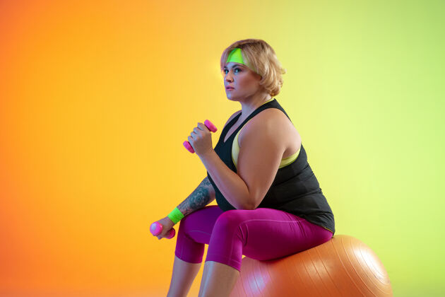 拉伸年轻的白人大号女模特在霓虹灯下的橙色渐变背景上训练用重量做锻炼运动的概念 健康的生活方式 积极的身体 平等手臂运动装人