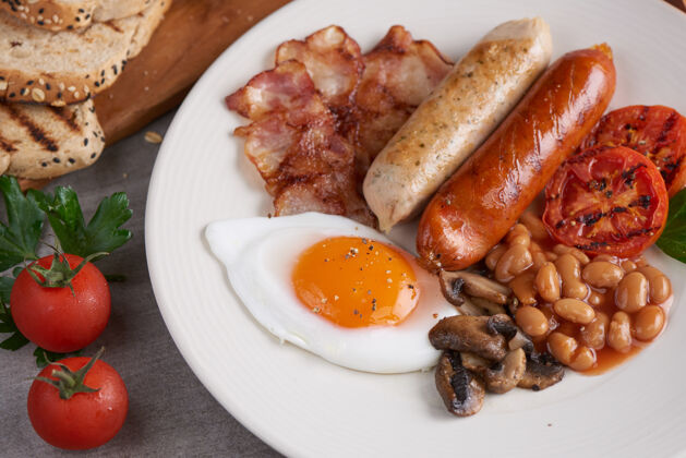 房子传统的全英式早餐 煎蛋 香肠 豆类 蘑菇 烤番茄和培根放在盘子里 吐司 黄油 果酱放在木板上美味丰盛面包