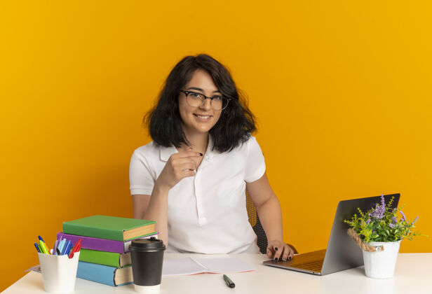 橙色年轻的笑容可掬的高加索女学生戴着眼镜坐在书桌旁 把学习工具孤立在橙色的空间里 还有复印空间桌子微笑工具
