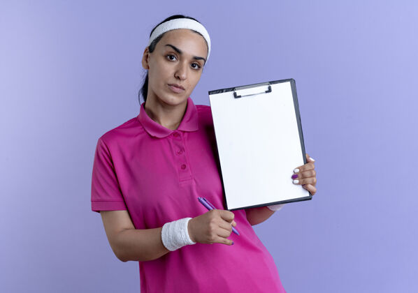 空间年轻自信的高加索运动女性戴着头带和腕带拿着笔和剪贴板孤立在紫色空间与复制空间运动剪贴板紫色