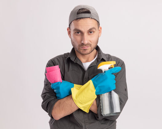 破布年轻的清洁工穿着休闲服 戴着橡胶手套 手里拿着清洁喷剂和抹布 站在橙色的墙上 表情严肃立场清洁衣服