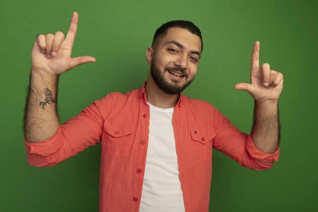 表演身穿橘色衬衫 留着胡子的年轻人面带微笑 食指站在绿色的墙上胡须微笑手指