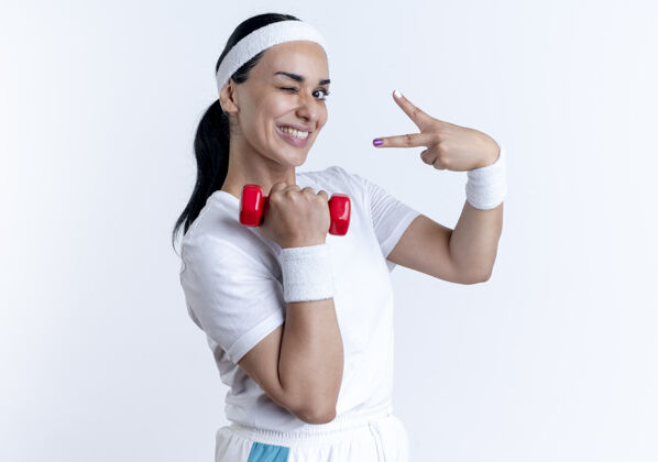 标志年轻微笑的白人运动女性戴着头带和腕带眨着眼睛举着哑铃和手势胜利的手标志隔离在白色空间与复制空间运动空间佩戴