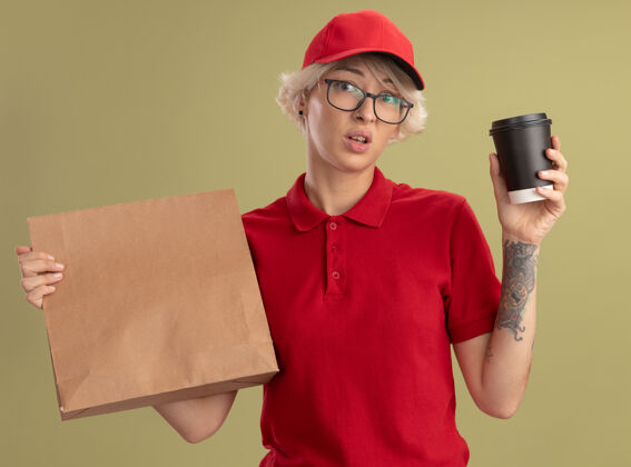 拿着年轻的送货员身穿红色制服 戴着帽子 戴着眼镜 手里拿着纸包和咖啡杯 困惑而焦急地站在绿色的墙上站着杯子帽子