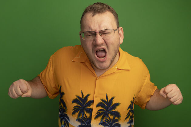 男人一个戴着眼镜 穿着橙色衬衫的男人站在绿色的墙上 握紧拳头大喊大叫 愤怒又沮丧站着沮丧喊叫