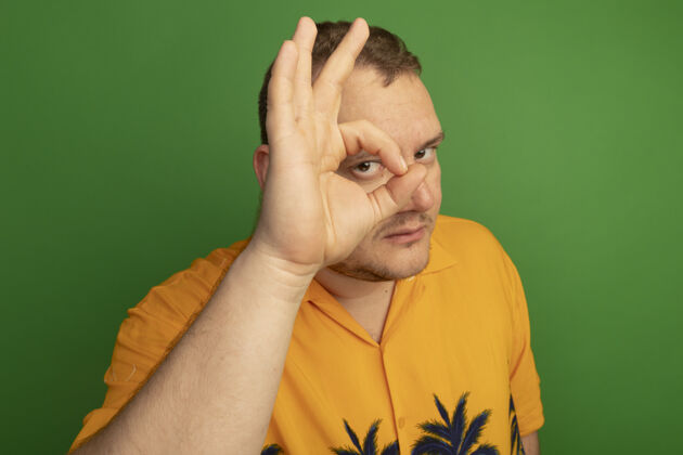 伙计一个戴着眼镜 穿着橘色衬衫的男人正在做一个“ok”的手势 他站在绿色的墙上 透过这个手势看着通过眼镜好的