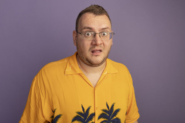 站着一个戴着眼镜 穿着橙色衬衫的男人站在紫色的墙上 困惑和担心男人担心戴着