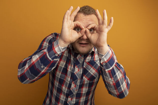 手指戴着眼镜和格子衬衫的男人站在橙色的墙上 用手指做着双眼的手势男人眼镜通过
