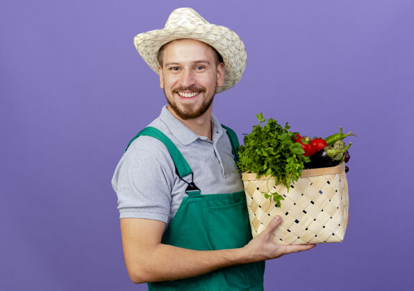篮子快乐的年轻帅气的斯拉夫园丁 穿着制服 戴着帽子 手里拿着一篮蔬菜 隔离在紫色的墙上 留着复制空间看着拿着斯拉夫人