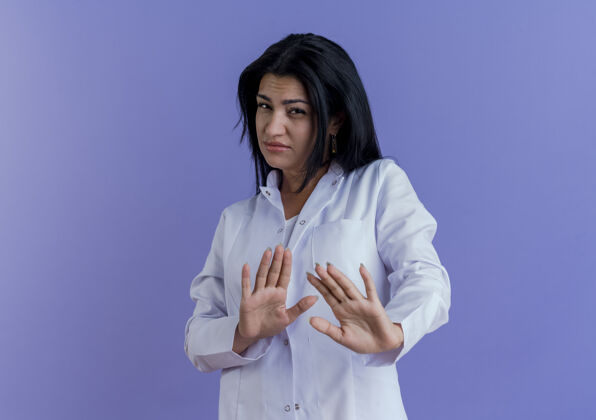表情未出租的年轻女医生穿着医疗袍做没有手势孤立在紫色的墙上与复制空间年轻女性医生