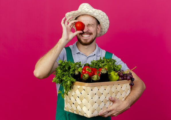 帽子面带微笑的年轻英俊的斯拉夫园丁 身着制服 戴着帽子 手里拿着一篮蔬菜 眼睛紧闭着西红柿 孤零零地站在深红色的墙上斯拉夫花园微笑