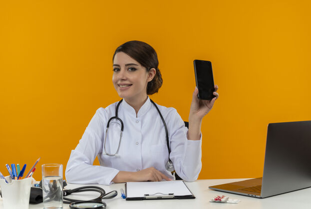 女性面带微笑的年轻女医生穿着医用长袍 听诊器坐在办公桌旁 手里拿着医疗工具和笔记本电脑 黄色的墙上贴着隔离的手机笔记本电脑工具办公桌