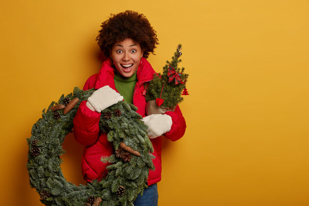 黑发惊讶的美国黑人妇女手持绿色花环和枞树 表情欢快 准备过圣诞节或新年 被隔离在黄色的墙壁上高兴节日站立