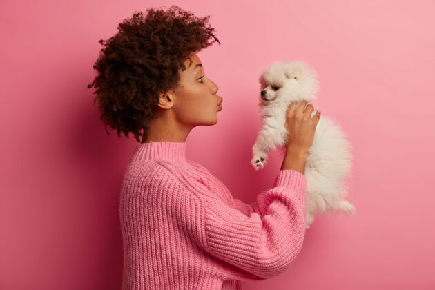 青少年美国黑人妇女亲吻繁殖的狗 举起手来 穿着针织毛衣 在粉色背景下摆姿势黑发狗亲吻