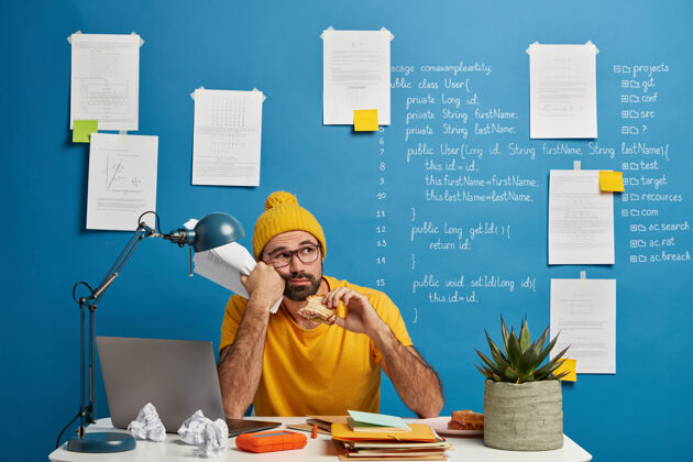 事业体贴的男性程序员或软件开发人员思考程序代码 把目光移开吃汉堡包 拿着文件 穿着黄色衣服 花时间做项目思想经理帽子