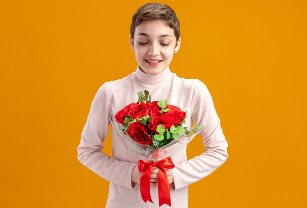 年轻一位留着短发的年轻女子手持一束红玫瑰 微笑着看着玫瑰 站在橘红色的墙上庆祝情人节短裤情人节玫瑰