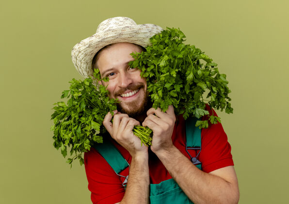 看面带微笑的年轻英俊的斯拉夫园丁 身着制服 戴着帽子 手拿香菜 脸贴在橄榄绿的墙上帽子近花园