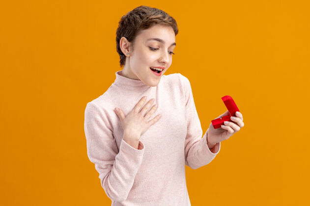 天一位留着短发的年轻女士拿着一个红色的盒子和一枚订婚戒指看着它 站在橘色的墙上 惊喜而快乐的情人节概念戒指立场概念