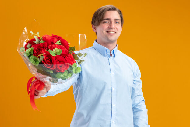 微笑身着蓝衣的快乐男人手持一束红玫瑰 看着相机 站在橙色的墙上 欢快地微笑着情人节的概念概念玫瑰日子
