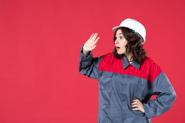 硬特写镜头中惊讶的女建筑工人穿着制服 戴着安全帽 专注于孤立的红墙上的东西成人快乐时尚