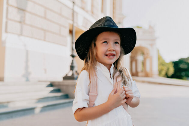 高兴可爱可爱的小女孩戴着帽子探索城市姿势金发背包