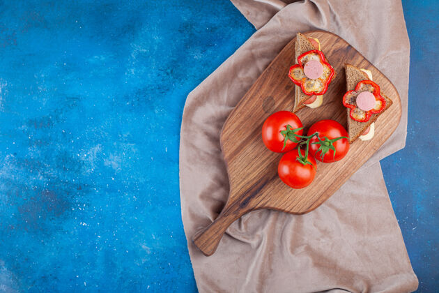 面包三明治上放着香肠和整个西红柿 放在切菜板上 蓝色的布料上酵母美味美味