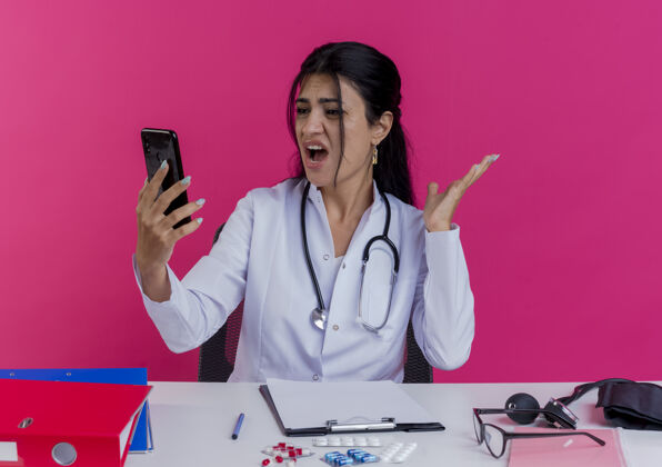 坐着愤怒的年轻女医生穿着医用长袍 手持听诊器坐在办公桌旁 手持医疗工具 看着手机 一只空手孤立在粉红色的墙上工具手机听诊器
