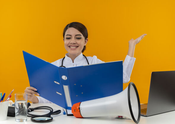 看着面带微笑的年轻女医生穿着医用长袍和听诊器坐在办公桌旁 手持医疗工具 扬声器和笔记本电脑 看着黄色墙上显示空手的文件夹笔记本电脑长袍坐着