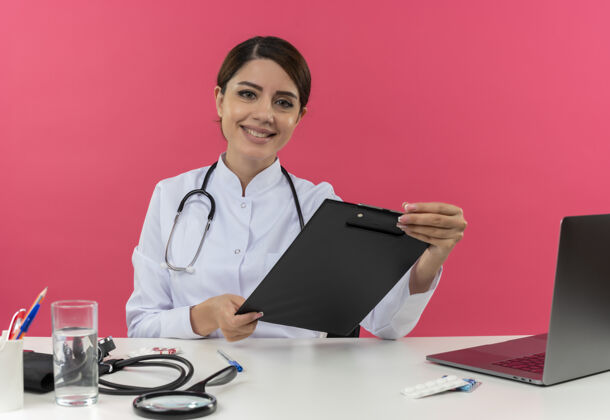 医生面带微笑的年轻女医生穿着医用长袍 听诊器坐在办公桌旁 手里拿着医疗工具和笔记本电脑 把剪贴板孤立地放在粉红色的墙上微笑穿办公桌