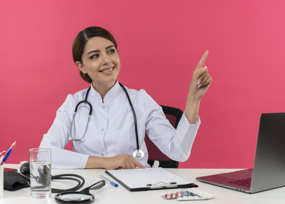 工具面带微笑的年轻女医生穿着医用长袍和听诊器坐在办公桌旁 手里拿着医疗工具和笔记本电脑 看着并指着隔离在粉红色墙上的一侧微笑听诊器指着