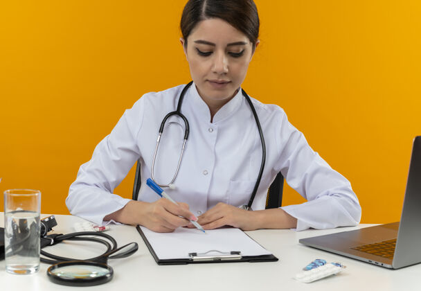 听诊器专注的年轻女医生穿着医用长袍和听诊器坐在办公桌旁 手里拿着医疗工具和笔记本电脑 写字板上的笔被隔离在黄色的墙上笔记本电脑医疗桌子