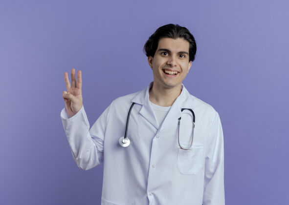 医疗快乐的年轻男医生穿着医用长袍和听诊器做和平的标志隔离在紫色的墙上复制空间长袍穿和平