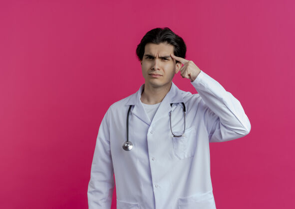 听诊器皱眉的年轻男医生穿着医用长袍和听诊器手指着太阳穴隔离在粉红色的墙上有复印空间男性医学手指