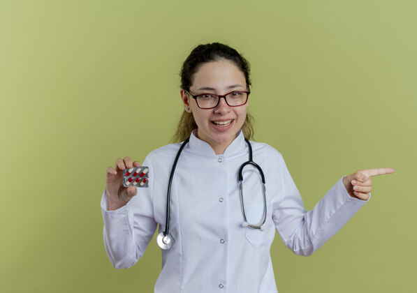 年轻面带微笑的年轻女医生 穿着医用长袍 戴着听诊器 戴着眼镜 拿着药片 指着隔离在橄榄绿墙上的有复印空间的一边医生眼镜女