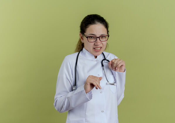 女神经质的年轻女医生 穿着医用长袍 戴着听诊器 橄榄绿墙上的眼镜戴吱吱声长袍