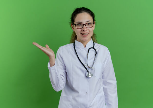 医生快乐的年轻女医生 穿着医用长袍 戴着听诊器和眼镜 在绿色的墙壁上展示着被隔离的空手和复印空间女长袍空